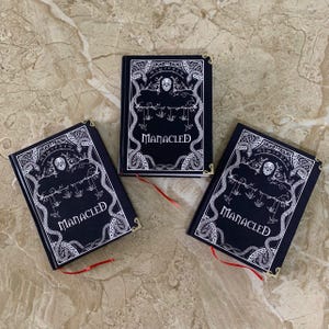 Pu&ograve; includere: Tre libri neri con copertina rigida con intricati disegni bianchi e la parola "MANACLED". Ogni libro ha un segnalibro a nastro rosso e angoli dorati. Le copertine presentano teschi, serpenti e elementi gotici.