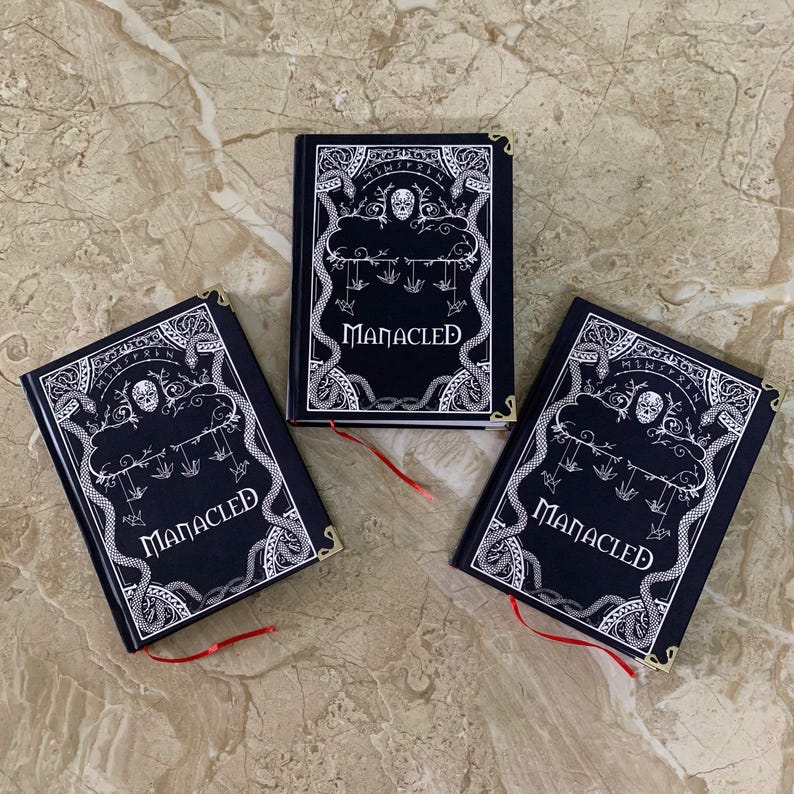 Pu&ograve; includere: Tre libri neri con copertina rigida con intricati disegni bianchi e la parola "MANACLED". Ogni libro ha un segnalibro a nastro rosso e angoli dorati. Le copertine presentano teschi, serpenti e elementi gotici.
