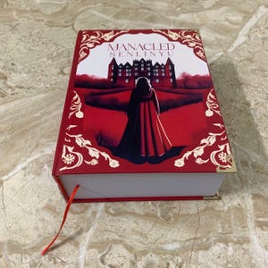 K&ouml;nnte beinhalten: Ein Hardcover-Buch mit rotem Einband, dem Titel "MANACLED SENLINYU" und einer Illustration einer Person in rotem Umhang vor einem Schloss. Das Buch hat goldene Buchecken und ein rotes Leseb&auml;ndchen.