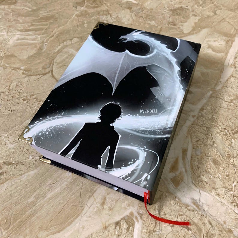 K&ouml;nnte beinhalten: Ein schwarz-wei&szlig; illustriertes Buch mit einem Drachen und einer Silhouette auf dem Einband. Das Buch hat goldfarbene Eckensch&uuml;tzer und ein rotes Leseb&auml;ndchen. Das Wort "AVENDELL" ist auf dem Einband aufgedruckt.