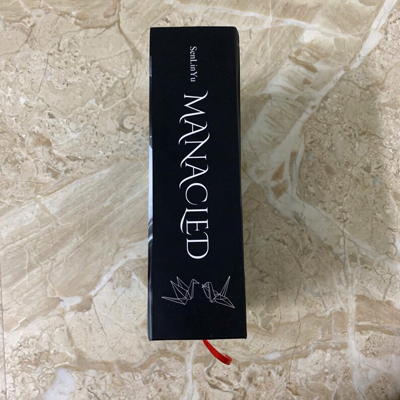 K&ouml;nnte beinhalten: Ein schwarzes Buch mit dem Titel "MANACIED" in wei&szlig;er Schrift. Der Name des Autors, SenLinYu, ist &uuml;ber dem Titel aufgedruckt. Zwei Origami-V&ouml;gel befinden sich am unteren Rand. Ein rotes Leseb&auml;ndchen ist sichtbar.