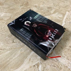 K&ouml;nnte beinhalten: Ein Hardcover-Buch mit dem Titel "MANACLED" mit einem dunklen Cover, das zwei Figuren zeigt. Das Buch hat ein rotes Leseb&auml;ndchen und goldene Eckakzente. Der Name des Autors, Sen Lin Yu, ist auf dem Cover zu sehen.