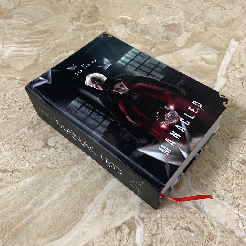 K&ouml;nnte beinhalten: Ein Hardcover-Buch mit dem Titel "MANACLED" mit einem dunklen Cover, das zwei Figuren zeigt. Das Buch hat ein rotes Leseb&auml;ndchen und goldene Eckakzente. Der Name des Autors, Sen Lin Yu, ist auf dem Cover zu sehen.