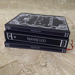 Pu&ograve; includere: Tre libri con copertina rigida neri impilati l'uno sull'altro. I libri sono intitolati "Manacled" in testo bianco. Il libro in alto ha un design bianco ornato. I libri hanno segnalibri a nastro rosso. I libri sono su una superficie marrone chiaro.