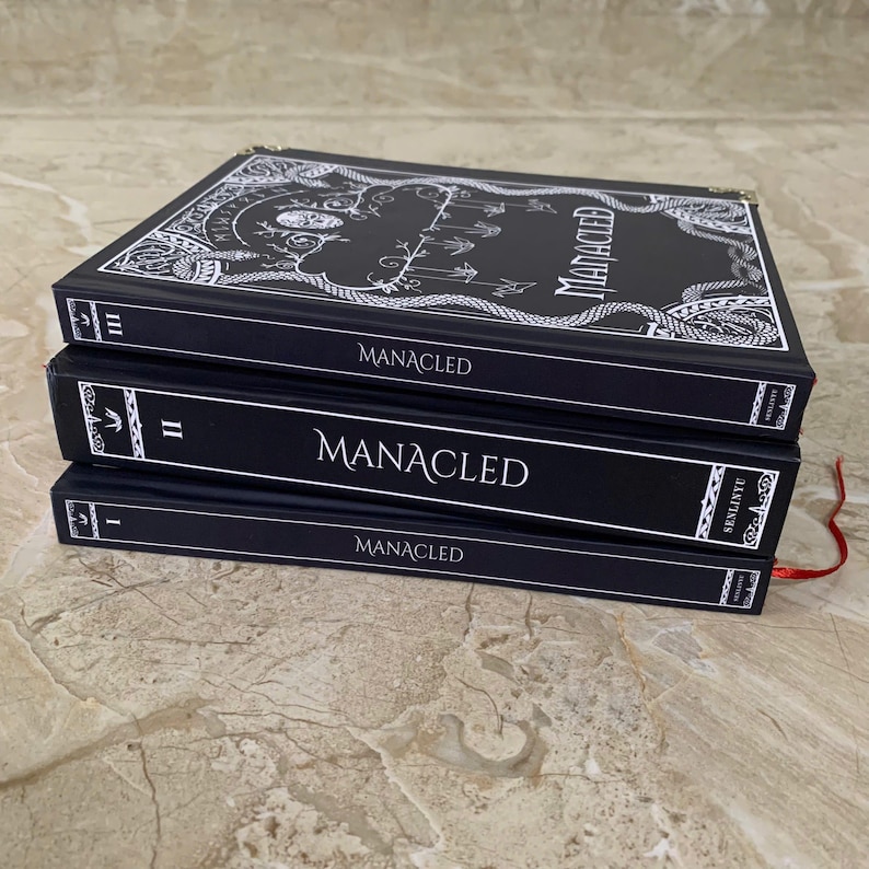 Pu&ograve; includere: Tre libri con copertina rigida neri impilati l'uno sull'altro. I libri sono intitolati "Manacled" in testo bianco. Il libro in alto ha un design bianco ornato. I libri hanno segnalibri a nastro rosso. I libri sono su una superficie marrone chiaro.