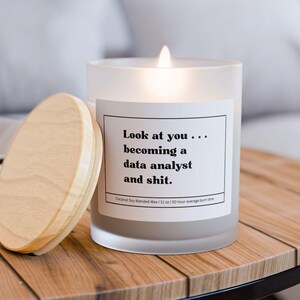 May include: A lit, frosted glass candle with a wooden lid on a wooden table. The candle has a white label with black text: "Look at you... becoming a data analyst and shit." The candle is next to its wooden lid.