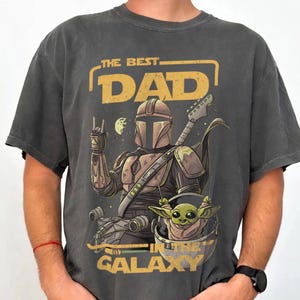 Pu&ograve; includere: T-shirt grigio scuro con una grafica raffigurante un personaggio in stile Mandaloriano che suona la chitarra, con Grogu in una capsula. Il testo recita "THE BEST DAD IN THE GALAXY" in giallo. La maglietta ha un aspetto vintage.