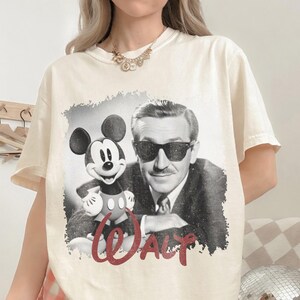 K&ouml;nnte beinhalten: Cremefarbenes T-Shirt mit einem Schwarz-Wei&szlig;-Bild von Mickey Mouse und Walt Disney. Das Wort "WALT" ist in roter Schrift darunter geschrieben. Das Shirt hat kurze &Auml;rmel und eine lockere Passform.