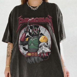 May include: Dark gray t-shirt featuring a graphic of Boba Fett holding a blaster and a Stormtrooper helmet. The design is set within a white circle with a distressed look, and the word "Mandalorian" is printed above the image in red. The shirt has short sleeves.