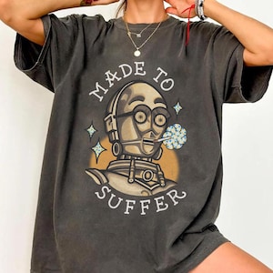Puede incluir: Camiseta gris oscuro con una ilustración de robot y el texto "MADE TO SUFFER". El robot fuma una flor azul y blanca. El diseño incluye un círculo amarillo y estrellas.