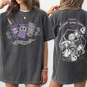 May include: Dark gray t-shirt featuring a graphic from Tim Burton's "The Nightmare Before Christmas." The front design shows three playing cards with skull faces and the text "3 of a Kind." The back displays a detailed scene with characters from the movie.