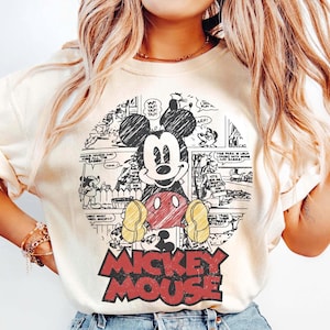 May include: Cream-colored sweatshirt featuring a Mickey Mouse graphic. The graphic includes a circular comic strip design with Mickey Mouse in the center, wearing red shorts and yellow shoes. The words "MICKEY MOUSE" are in red block letters at the bottom.