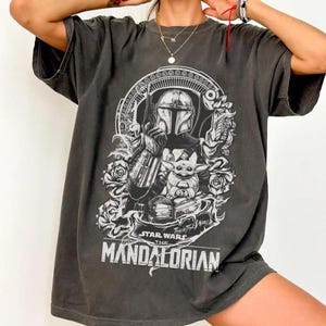 May include: Dark gray t-shirt featuring a graphic of The Mandalorian and Grogu from Star Wars. The design includes the text "Star Wars The Mandalorian" and "This is the Way" in white. The shirt has a crew neck and short sleeves.