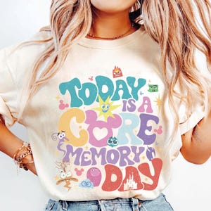 May include: A cream-coloured sweatshirt featuring the phrase "Today is a Core Memory Day" in a vibrant, retro font. The design incorporates cartoon characters and a castle illustration, creating a whimsical and nostalgic feel.