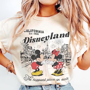 May include: Cream-colored Disneyland sweatshirt featuring Mickey and Minnie Mouse in a vintage design. The graphic includes the words "California Est. 1955" and "Disneyland" with fireworks and a cityscape. The phrase "The happiest place on earth" is also included.