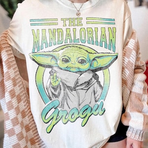 May include: Cream-colored t-shirt featuring a graphic of Grogu from The Mandalorian. The design includes the text "THE MANDALORIAN" and "Grogu" in green and blue. The image of Grogu is in the center, surrounded by a blue and green circle.