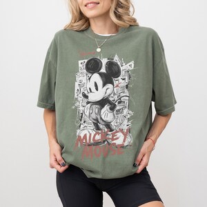May include: Olive green t-shirt featuring a graphic of Mickey Mouse in a black and white graffiti style. The words "Mickey Mouse" are printed in red at the bottom. The shirt has short sleeves and a relaxed fit.