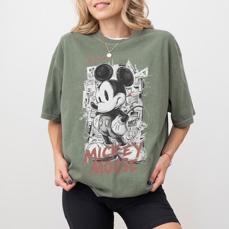 May include: Olive green t-shirt featuring a graphic of Mickey Mouse in a black and white graffiti style. The words "Mickey Mouse" are printed in red at the bottom. The shirt has short sleeves and a relaxed fit.