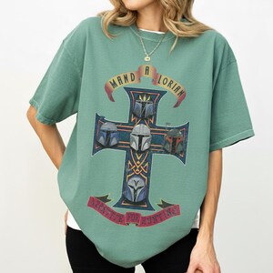 以下が含まれることがあります： セージグリーンの半袖Tシャツ。4つの異なるマンダロリアンのヘルメットが描かれた十字架の大きなグラフィックが特徴です。十字架の上部のバナーには「MAND A LORIAN」、下部のバナーには「APPETITE FOR HUNTING」という文字がプリントされています。
