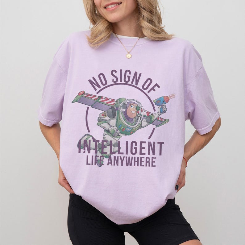 Op de afbeelding: Lichtpaarse t-shirt met een afbeelding van Buzz Lightyear en de tekst "NO SIGN OF INTELLIGENT LIFE ANYWHERE". Het ontwerp toont Buzz in zijn ruimtepak, met een straalpistool en vliegend.