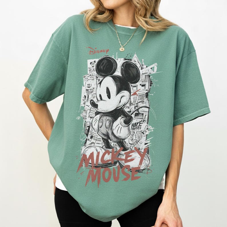 May include: Sage green t-shirt featuring a graphic of Mickey Mouse in black, white, and red. The design includes the Disney logo and the words "Mickey Mouse" in a distressed font. The shirt has short sleeves and a relaxed fit.
