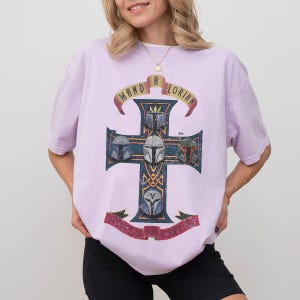 以下が含まれることがあります： ライトパープルの半袖Tシャツ。5つのマンダロリアン・ヘルメットが描かれたダークブルーの十字架の大きなグラフィックが特徴です。十字架の上には黄色いバナーに「MANDALORIAN」の文字がアーチ状に配置され、下部の赤いバナーにも文字が記されています。