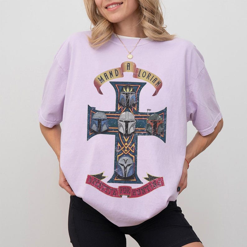 以下が含まれることがあります： ライトパープルの半袖Tシャツ。5つのマンダロリアン・ヘルメットが描かれたダークブルーの十字架の大きなグラフィックが特徴です。十字架の上には黄色いバナーに「MANDALORIAN」の文字がアーチ状に配置され、下部の赤いバナーにも文字が記されています。