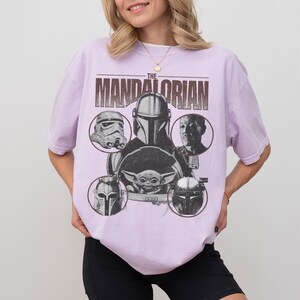 Pu&ograve; includere: Una maglietta lilla con una grafica dei personaggi di "The Mandalorian". Il design include il testo "THE MANDALORIAN" sopra immagini circolari di personaggi come il Mandaloriano, uno Stormtrooper e Grogu.