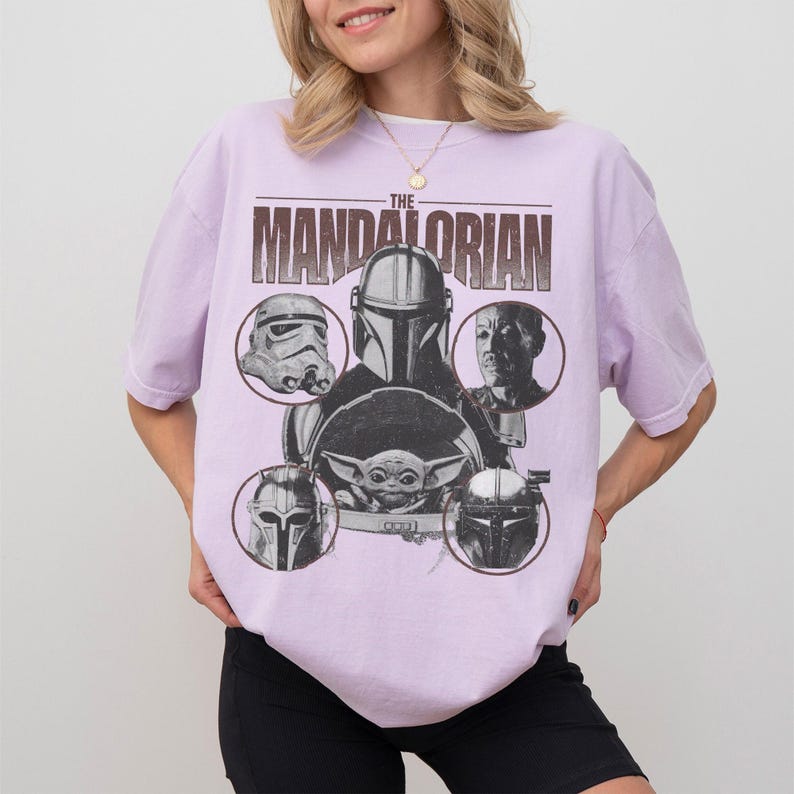 Pu&ograve; includere: Una maglietta lilla con una grafica dei personaggi di "The Mandalorian". Il design include il testo "THE MANDALORIAN" sopra immagini circolari di personaggi come il Mandaloriano, uno Stormtrooper e Grogu.