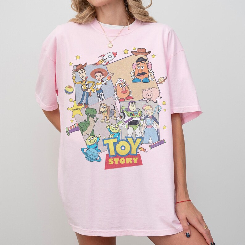 Puede incluir: Camiseta rosa claro con un gr&aacute;fico colorido de personajes de la pel&iacute;cula animada "Toy Story". El dise&ntilde;o incluye a Woody, Buzz Lightyear y otros personajes, con las palabras "TOY STORY" en una fuente audaz y colorida.