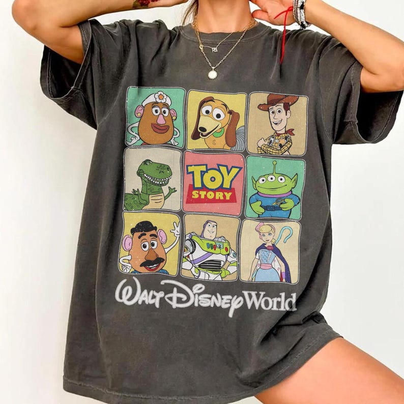 May include: Dark gray t-shirt featuring a colorful grid of Toy Story characters, including Mr. Potato Head, Slinky Dog, Woody, Rex, and Buzz Lightyear. The shirt also has the words "Toy Story" and "Walt Disney World" printed on it.