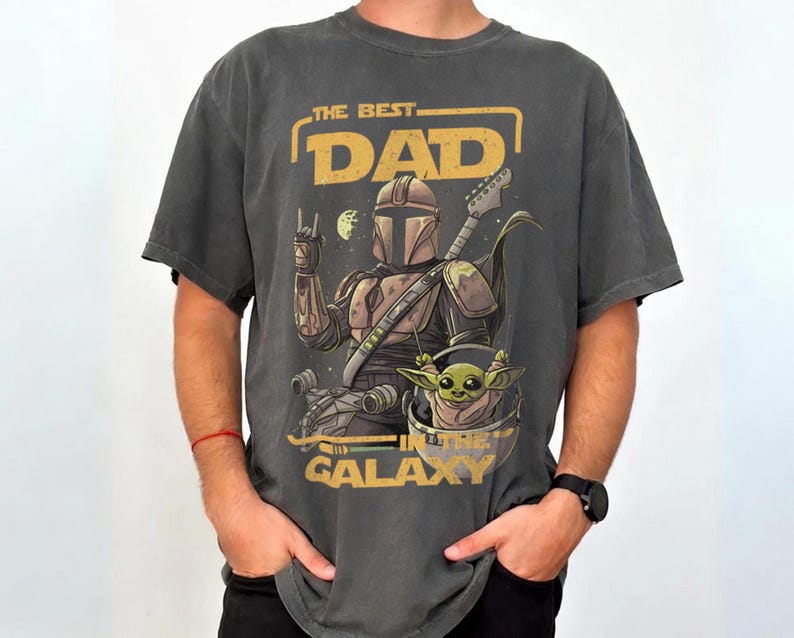 Pu&ograve; includere: T-shirt grigio scuro con una grafica raffigurante un personaggio in stile Mandaloriano che suona la chitarra, con Grogu in una capsula. Il testo recita "THE BEST DAD IN THE GALAXY" in giallo. La maglietta ha un aspetto vintage.