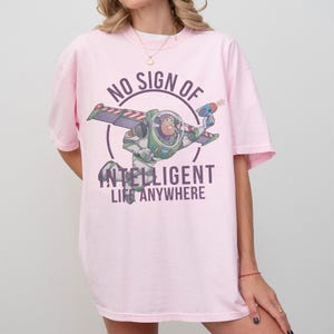 Op de afbeelding: Een lichtroze t-shirt met een afbeelding van Buzz Lightyear uit Toy Story. Het ontwerp bevat de tekst "NO SIGN OF INTELLIGENT LIFE ANYWHERE" in een cirkelvormige lay-out rond het personage. Het shirt heeft korte mouwen.