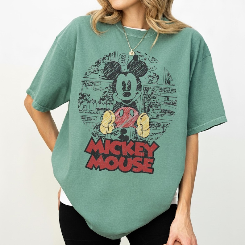 May include: Green t-shirt featuring a graphic of Mickey Mouse in a comic book style. The design includes Mickey's face, red shorts, yellow shoes, and the words "Mickey Mouse" in red. The shirt has short sleeves and a crew neck.