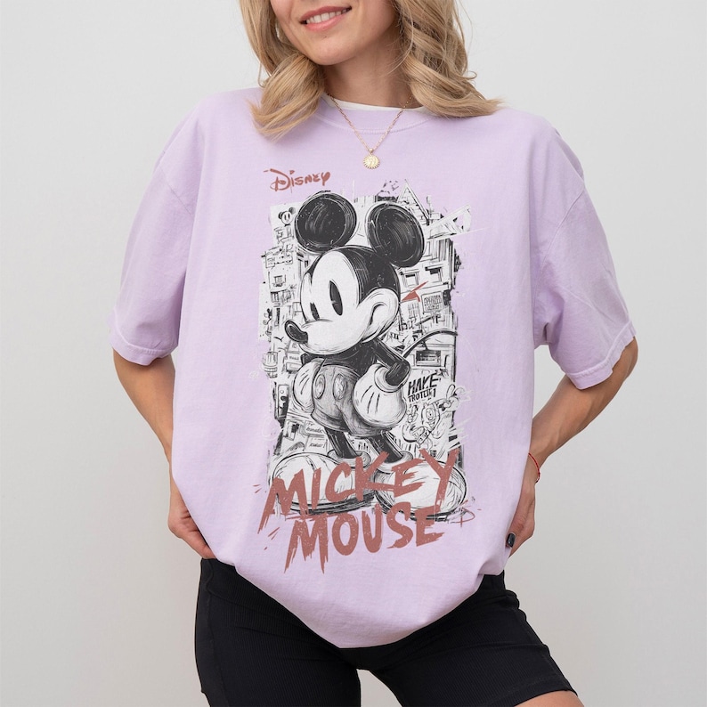 May include: A light purple t-shirt featuring a black and white graphic of Mickey Mouse. The design includes the word "Disney" and "Mickey Mouse" in a stylized font. The shirt has short sleeves and a relaxed fit.