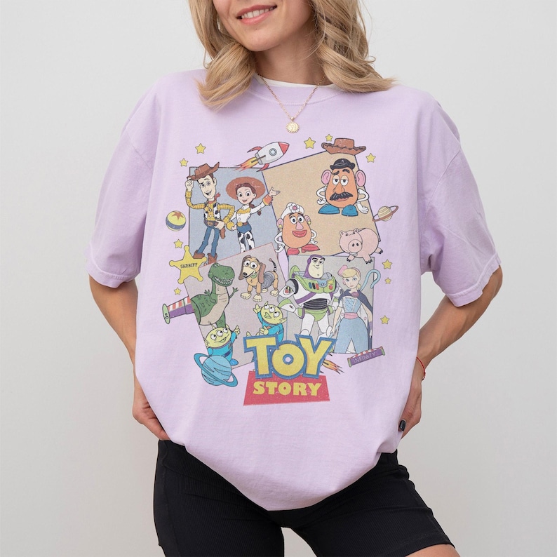 Puede incluir: Una camiseta de color lila claro con un gr&aacute;fico colorido de personajes de la pel&iacute;cula animada "Toy Story". El dise&ntilde;o incluye a Woody, Buzz Lightyear y otros personajes, con las palabras "Toy Story" en letras grandes.