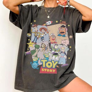 Puede incluir: Camiseta gris oscuro con un gr&aacute;fico colorido de personajes de la pel&iacute;cula animada "Toy Story". El dise&ntilde;o incluye a Woody, Buzz Lightyear y otros personajes, con las palabras "Toy Story" en la parte inferior. La camiseta tiene un aspecto vintage y desgastado.