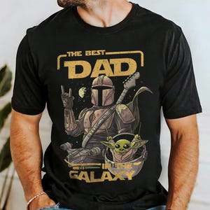 Pu&ograve; includere: T-shirt nero con una grafica raffigurante un personaggio in stile Mandaloriano che suona la chitarra, con la scritta "THE BEST DAD IN THE GALAXY". Il disegno include un piccolo alieno verde. La maglietta &egrave; girocollo.