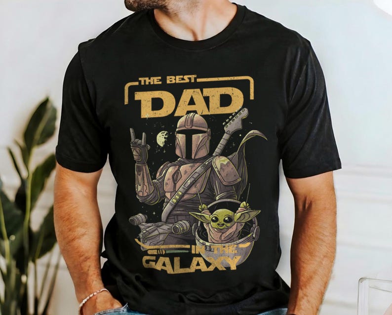 Pu&ograve; includere: T-shirt nero con una grafica raffigurante un personaggio in stile Mandaloriano che suona la chitarra, con la scritta "THE BEST DAD IN THE GALAXY". Il disegno include un piccolo alieno verde. La maglietta &egrave; girocollo.