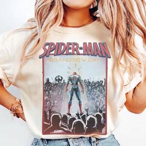May include: Cream-coloured sweatshirt with a Spider-Man graphic. The graphic features the words "SPIDER-MAN" in red and blue, with "BRAND NEW DAY" below. The image shows Spider-Man standing before a crowd of people.