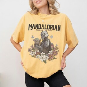 Op de afbeelding: Mosterdgeel T-shirt met een afbeelding van The Mandalorian en Grogu, omringd door bloemen en vlinders. De tekst "MANDALORIAN - THIS IS THE WAY" staat in het zwart gedrukt. Het T-shirt heeft een ronde hals en korte mouwen.