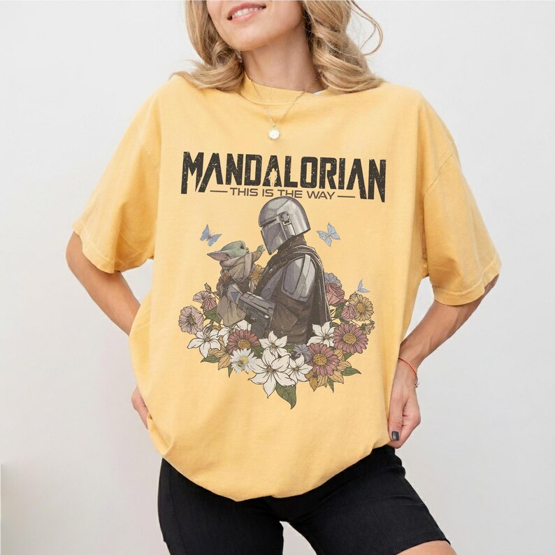 Op de afbeelding: Mosterdgeel T-shirt met een afbeelding van The Mandalorian en Grogu, omringd door bloemen en vlinders. De tekst "MANDALORIAN - THIS IS THE WAY" staat in het zwart gedrukt. Het T-shirt heeft een ronde hals en korte mouwen.