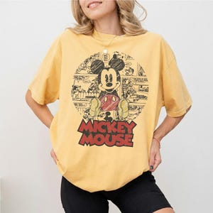 May include: Mustard yellow t-shirt featuring a graphic of Mickey Mouse in a comic strip style. The design includes Mickey Mouse in a seated pose with a red shorts and yellow shoes. The words "Mickey Mouse" are printed in red block letters below the image.
