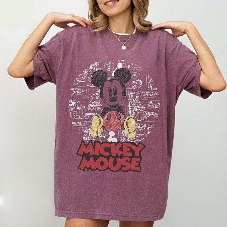 May include: A maroon t-shirt featuring a graphic of Mickey Mouse. The design includes a circular comic strip background and the words "Mickey Mouse" in bold, red and black letters. The shirt has short sleeves.