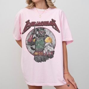 May include: A light pink t-shirt featuring a graphic of Boba Fett from Star Wars. The design includes the word "BAMALORIAN" in a distressed, metallic font above the image of the character. The graphic is set against a circular, distressed background.