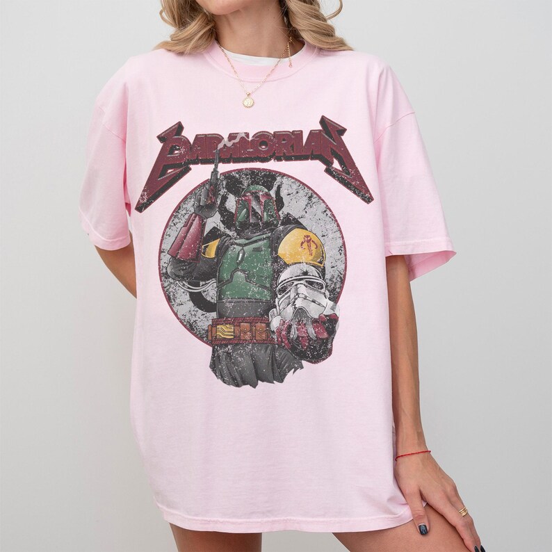 May include: A light pink t-shirt featuring a graphic of Boba Fett from Star Wars. The design includes the word "BAMALORIAN" in a distressed, metallic font above the image of the character. The graphic is set against a circular, distressed background.