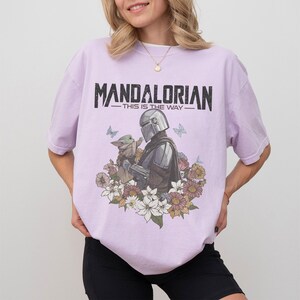 Op de afbeelding: Lichtpaars T-shirt met een afbeelding van The Mandalorian en Grogu, omringd door bloemen en vlinders. De tekst "MANDALORIAN - THIS IS THE WAY" staat in zwart bovenaan.