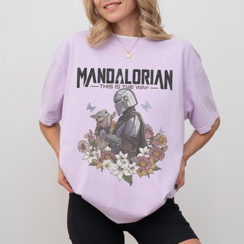Op de afbeelding: Lichtpaars T-shirt met een afbeelding van The Mandalorian en Grogu, omringd door bloemen en vlinders. De tekst "MANDALORIAN - THIS IS THE WAY" staat in zwart bovenaan.