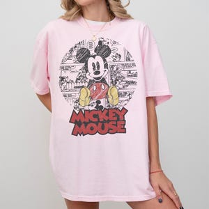 May include: A light pink t-shirt featuring a large graphic of Mickey Mouse. The graphic includes a black and white comic strip background and the words "Mickey Mouse" in red and black. The shirt has short sleeves and a relaxed fit.