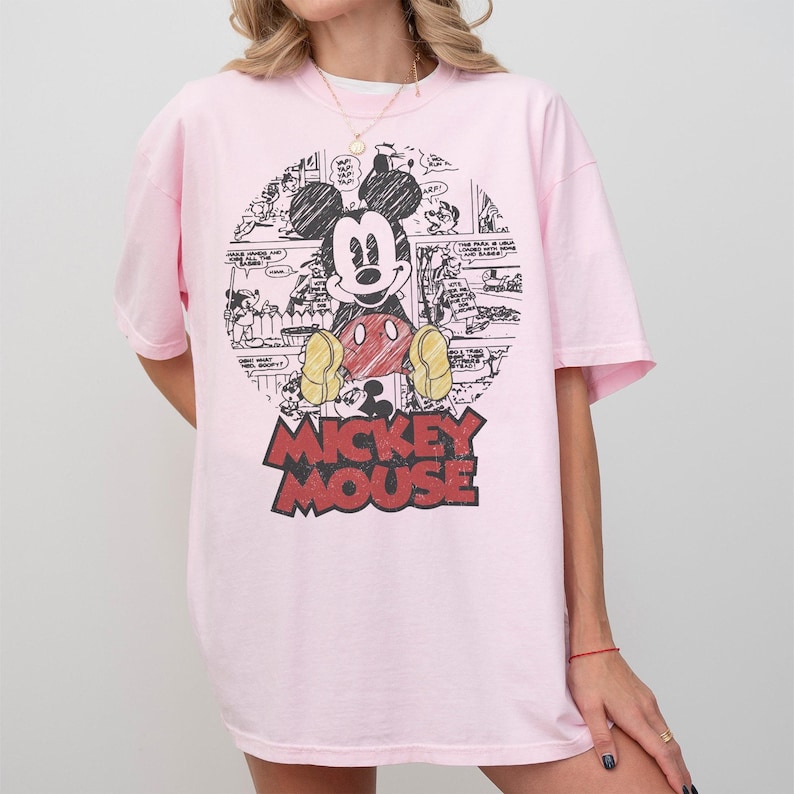 May include: A light pink t-shirt featuring a large graphic of Mickey Mouse. The graphic includes a black and white comic strip background and the words "Mickey Mouse" in red and black. The shirt has short sleeves and a relaxed fit.